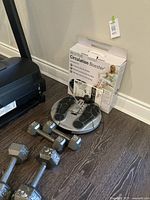 Revitive Circulation Booster product box and device with foot pads visible, along with four metal hex dumbbell weights (two labeled 10 lb and two labeled 20 lb) placed on the floor on wood surface.