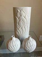Set of three white bisque porcelain vases, 1 large and 2 small, with raised floral patterns displayed on glass surface.