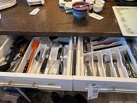 Open kitchen drawers showing stainless steel cutlery organized in plastic trays and various kitchen utensils including ladles, peelers, and spatulas.
