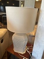White ceramic table lamp sitting on a wood side table, showing full lamp including base and cylindrical white shade.