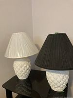 Photo showing both white ceramic table lamps side by side on a black table, one with a black pleated shade and one with a white shade covered in plastic.