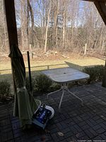 Patio table with folded green umbrella and box of patio lights on a brick patio with background of lawn and trees