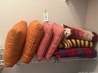 Full set of four throw pillows (two orange, two pink) and three blankets (two knitted multicolor, one plaid) stacked on a shelf.