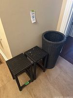 Photo showing two black wicker square plant stands and one tapered cylindrical wicker planter against a wall on a tiled floor.