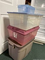 Stack of four Rubbermaid plastic storage totes: small clear blue tote, large clear tote with gray lid, medium-size clear pink Roughneck tote, and large beige Roughneck tote with brown lid