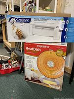 Photo shows boxed Garrison Plug-In Convection Heater on top of boxed Presto HeatDish parabolic electric heater, placed on floor with some shelving in background.