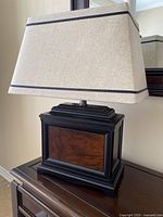 Front and angled views of a table lamp showing wood grain resin base with black framing and a beige fabric flared rectangular shade with dark trim.