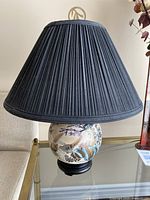 Full view showing black flared pleated fabric lampshade on a round porcelain hand-painted base with Asian motifs.