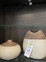 Two ceramic vases on a glass shelf, round and oval shapes with cream crackle finish and golden brown tops