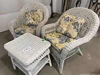 Two white wicker chairs with floral yellow and blue cushions and one white wicker side table shown together in garage setting.