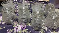 Photo showing multiple clear pressed glass punch cups with detailed embossed floral and geometric patterns, arranged upside down and right side up on a floral cloth.