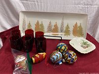 View of long rectangular ceramic platter featuring stylized Christmas trees, six deep red roly poly juice glasses, three colorful painted wooden ornaments, a fabric Santa ornament, and Mikasa trinket dish.
