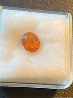 Oval orange-yellow faceted citrine gemstone resting on white cushioning in clear display box.