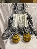 Full view of black and white paisley full kitchen apron with gathered waist and two small yellow glazed clay canisters with metal wire trim near apron base.