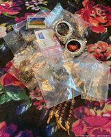Mixed assortment of costume jewelry laid on a floral fabric background, with large silver-tone clip-on earrings, pearl-like necklaces, wooden bead bracelets, and other assorted pieces in plastic bags.