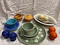 Full view of variety of ceramic and pottery items including cups, saucers, plates, serving dishes, and salt and pepper shakers on floral fabric background.