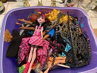 Purple container filled with multiple Monster High dolls laid together, showing various hair colors and a doll in a pink and black dress on top.