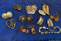 Photo shows a collection of vintage jewelry on blue fabric including pearl clip earrings, flower-shaped rhinestone earrings, gold-tone leaf brooch, heart-shaped brooch, and some rhinestone earrings and bracelet.