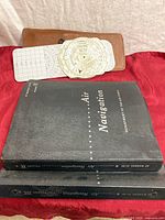Two black hardcover Air Navigation manuals stacked on each other, with visible titles and volume numbers, alongside the dead reckoning computer, grid charts, and leather case arranged above on red fabric.