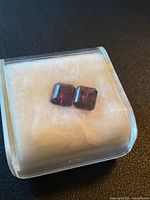 Pair of deep purple emerald cut amethyst stones shown on white fabric inside plastic box.