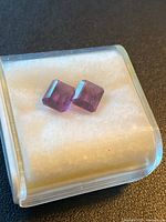 Pair of purple emerald cut Amethyst stones in a small clear box with white soft padding.