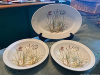 Two 10-inch rimmed serving bowls and one 14-inch oval serving platter displayed on a table, showing the floral botanical pattern in pastel colors on white porcelain.