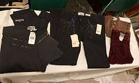 Overall view of five pairs of jeans and leggings laid on a table. Visible tags on most garments. Colors include black, brown, and burgundy.
