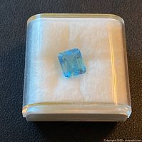 Photo of the loose emerald cut Swiss blue topaz gemstone in a small transparent display case, showing its vivid blue color and clear facets.