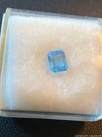 Single emerald cut blue topaz gemstone in a display case on white cotton background, highlighting the stone's color and cut.