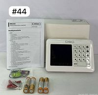 DSC WK250 keypad with backlit LCD, numeric keypad, included user manual, four AA batteries, two wrist key fobs, screws, and anchors arranged near keypad and box.