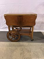 Side view of solid wood antique tea cart showing one large wooden decorative wheel and small caster wheels, lower storage shelf, and wing in folded position.