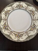 Close-up image of a dinner plate showing intricate green flower pattern on ivory/cream background with gold trim around the edge