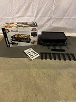 Full view showing one Swissmar raclette grill, 8 spatulas, 8 raclette dishes, instruction manual, and original box