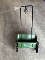 Front view of green Scotts AccuGreen 1000 spreader with black handle and wheels on concrete floor.