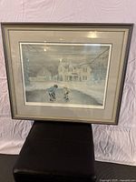 Front view of the framed print showing a winter scene with two children playing hockey on an icy road in front of houses.
