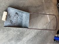 Top view of steel snow mover showing metal scoop, tubular handle, and stone weight on a concrete floor.