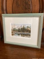 Framed print showing the landscape artwork with trees, water, and mountains, situated against a wooden chair background.