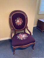 Side view of antique chair showing wooden curved armrests and legs, deep purple floral embroidered upholstery on backrest and seat.
