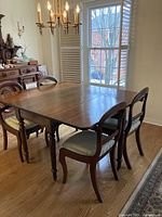 Dining area setup showing antique wooden gate leg table with six chairs around it, table top smooth and polished, chairs upholstered with light colored fabric seats.