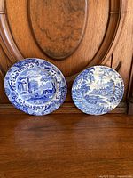 Two blue and white plates displayed upright on wooden surface and wood panel background, one larger with detailed ruins scene and floral edge, one smaller with rural scene.