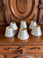 Six small ivory satin lamp shades arranged in two rows on wooden furniture, showing consistent shape and trim detail.