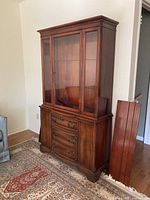 Front and side view of the wooden china cabinet standing on a patterned carpet. Shows glass doors at top with shelves and lower drawers and cupboard.