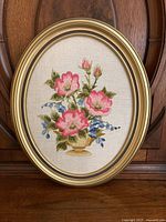 Oval crewel embroidery picture with pink and blue floral bouquet in yellow vase framed in gold-colored oval frame, shown against wood background.