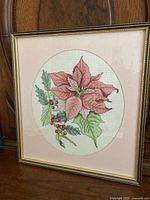 Framed cross stitch poinsettia artwork showing detailed stitch work and colors in a gold-tone frame with an oval mat.