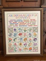 Full view of framed cross stitch sampler showing alphabet, flower names, and grid of floral motifs with initials, framed in wood.