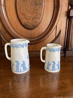 Front view of two Wedgwood pitchers, showing white and blue classical relief decoration and scale comparison.