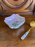 View of the pink floral square serving dish with scalloped edges and gold trim alongside the matching ladle on a wooden surface.