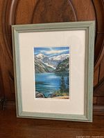 Framed watercolour print showing Lake Louise scene with mountains and water, in light wood or painted frame.