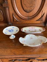Three china dishes displayed on wooden surface: blue floral pedestal dish, pink floral Dresden dish, and Silesia oval dish with hairline crack.