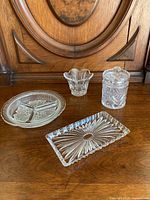 Four pressed glass dishes arranged on a wooden surface with a wood backdrop to show full lot contents including round divided dish, small decorative dish, jar with lid, and rectangular dish.